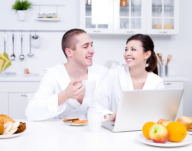 Felicidad de la joven pareja amorosa sentada en la cocina con el portátil