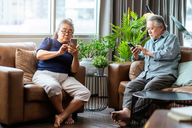 Felicidad pareja jubilada asiática disfruta jugando y compitiendo juego smartphone móvil en línea juntos en el sofá en la sala de estar casa interior backgroundasia pareja jugando juntos casa aislar idea