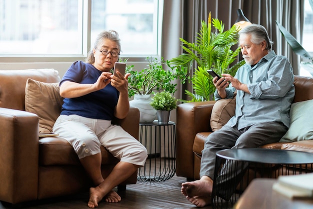 Felicidad pareja jubilada asiática disfruta jugando y compitiendo juego smartphone móvil en línea juntos en el sofá en la sala de estar casa interior backgroundasia pareja jugando juntos casa aislar idea