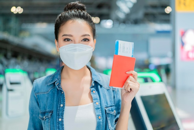 Felicidad sonriente mujer adulta asiática usar máscara facial gesto de la mano que muestra el pasaporte y el teléfono inteligente de pantalla en blanco en la terminal del aeropuerto mujer asiática lista para viajar después del cierre en el aeropuerto
