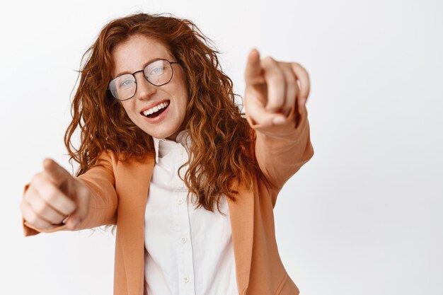 Felicidades Mujer de negocios pelirroja feliz con gafas señalando con el dedo a la cámara eligiendo que te recluten para unirte a la empresa de pie en traje contra fondo blanco