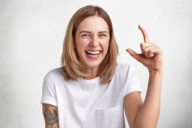 Feliz adorable joven con peinado bobbed muestra el tamaño de algo no muy grande, tiene una amplia sonrisa, muestra dientes blancos, vestida de manera informal