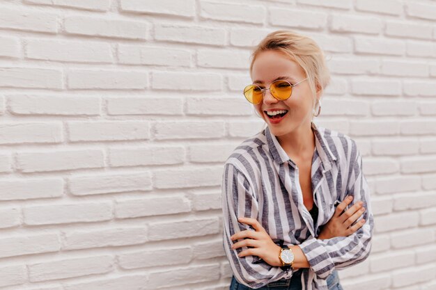 Feliz adorable mujer rubia con elegantes gafas naranjas en camisa a rayas posando con una sonrisa encantadora