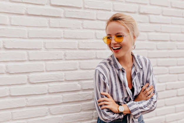 Feliz adorable mujer rubia con elegantes gafas naranjas en camisa a rayas posando con una sonrisa encantadora