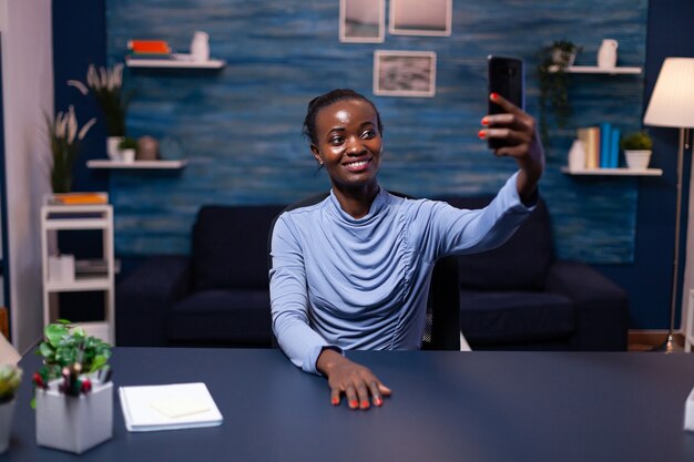 Feliz africano mirando smartphone tomando selfie mirando a cámara frontal. Trabajador autónomo ocupado que utiliza la red de tecnología moderna inalámbrica haciendo horas extraordinarias.