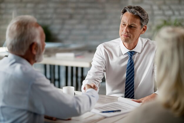 Feliz agente inmobiliario y clientes senior dándose la mano después de una reunión exitosa en la oficina