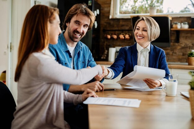 Feliz agente inmobiliario saludando a una pareja joven en una reunión en su casa. Las mujeres se dan la mano.