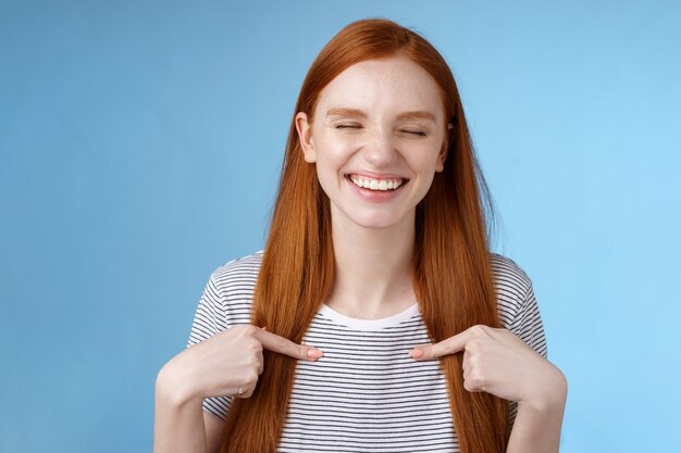 Feliz alegre alegre joven deportista pelirroja nominada sonriendo sorprendida riendo con alegría cerrar los ojos apuntando a sí misma elegida ganó el primer premio, de pie fondo azul