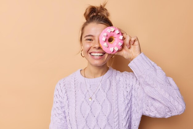 Feliz, alegre y despreocupada joven europea se cubre los ojos con un donut glaseado que va a comer un delicioso postre, lleva sonrisas de suéter de punto y posa ampliamente sobre un fondo beige. Concepto goloso