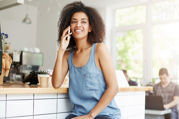 Feliz alegre hermosa mujer africana sonriente hablando por teléfono sentado en la cafetería.