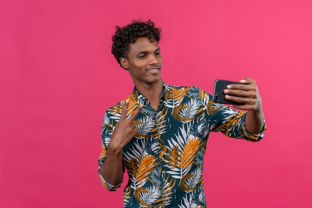 Feliz y alegre joven guapo de piel oscura con cabello rizado en hojas camisa estampada haciendo selfie con teléfono móvil mostrando dos dedos gesto