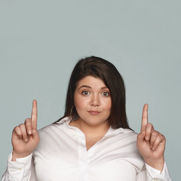 Feliz alegre joven mujer de pelo oscuro con mejillas regordetas y cuerpo grande con curvas mirando con mirada emocionada, apuntando con ambos dedos hacia arriba