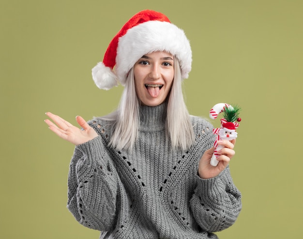 Feliz y alegre joven mujer rubia en suéter de invierno y gorro de Papá Noel con bastón de caramelo de navidad sacando la lengua de pie sobre la pared verde