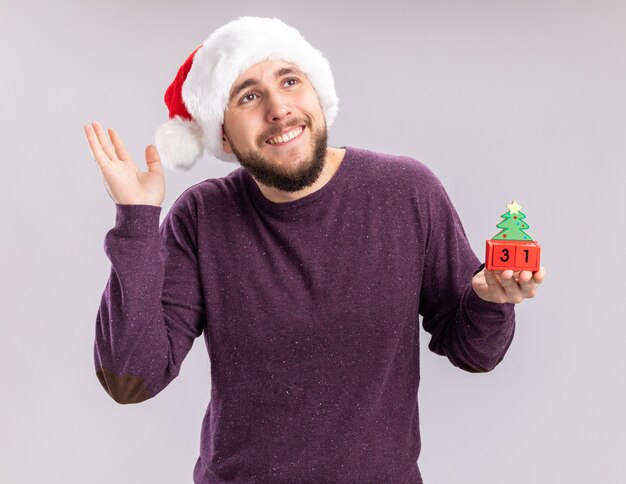 Feliz y alegre joven en suéter púrpura y gorro de Papá Noel mostrando cubos con fecha de año nuevo sonriendo alegremente de pie sobre fondo blanco.