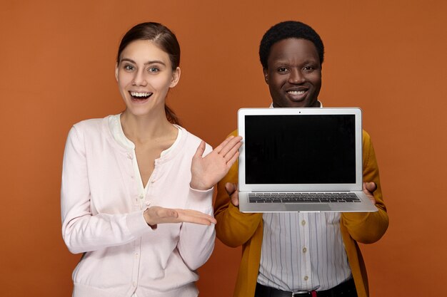 Feliz alegre joven vendedor afroamericano y su colega de aspecto amistoso emocionado de pie uno al lado del otro, anunciando un nuevo dispositivo electrónico portátil genérico y sonriendo