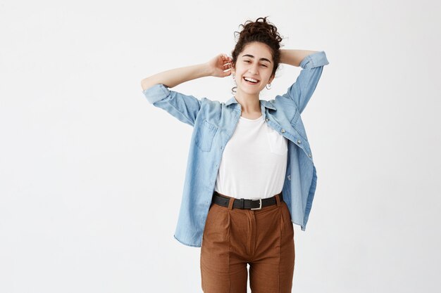 Feliz alegre joven vistiendo su cabello oscuro en bollo regocijándose en noticias positivas o regalo de cumpleaños, alegre y encantadoramente sonriendo. Chica estudiante positiva relajante en el interior después de la universidad