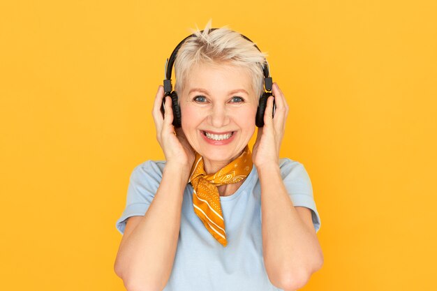 Feliz alegre mujer madura de pelo corto sonriendo ampliamente posando en amarillo en auriculares inalámbricos