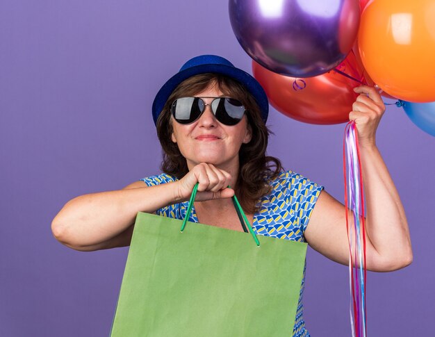 Feliz y alegre mujer de mediana edad con gorro de fiesta y gafas sosteniendo un montón de globos de colores y bolsas de papel con regalos celebrando la fiesta de cumpleaños de pie sobre la pared púrpura