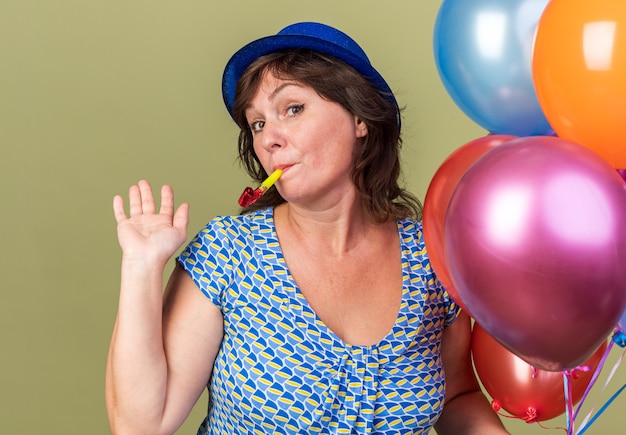 Feliz y alegre mujer de mediana edad con gorro de fiesta con un montón de globos de colores que sopla un silbato