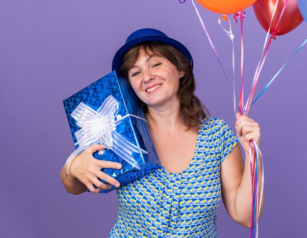 Feliz y alegre mujer de mediana edad con gorro de fiesta sosteniendo un montón de globos de colores y presente sonriendo ampliamente