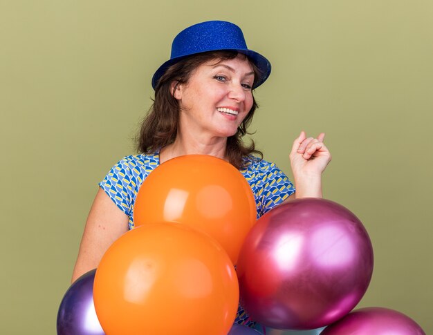 Feliz y alegre mujer de mediana edad con gorro de fiesta sosteniendo un montón de globos de colores sonriendo ampliamente celebrando la fiesta de cumpleaños de pie sobre la pared verde