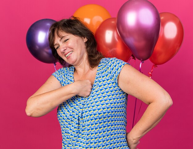 Feliz y alegre mujer de mediana edad con sombrero de fiesta con globos de colores sonriendo ampliamente