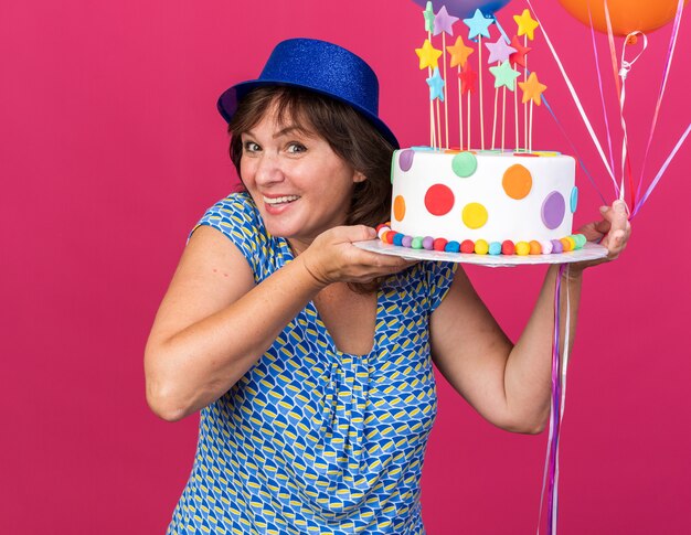 Feliz y alegre mujer de mediana edad con sombrero de fiesta con globos de colores sosteniendo pastel de cumpleaños sonriendo ampliamente celebrando la fiesta de cumpleaños de pie sobre la pared rosa