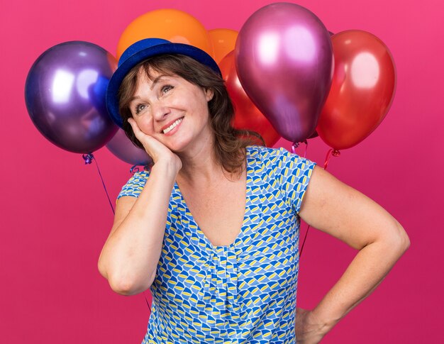 Feliz y alegre mujer de mediana edad con sombrero de fiesta sosteniendo globos de colores mirando hacia arriba con una sonrisa en la cara celebrando la fiesta de cumpleaños de pie sobre la pared rosa