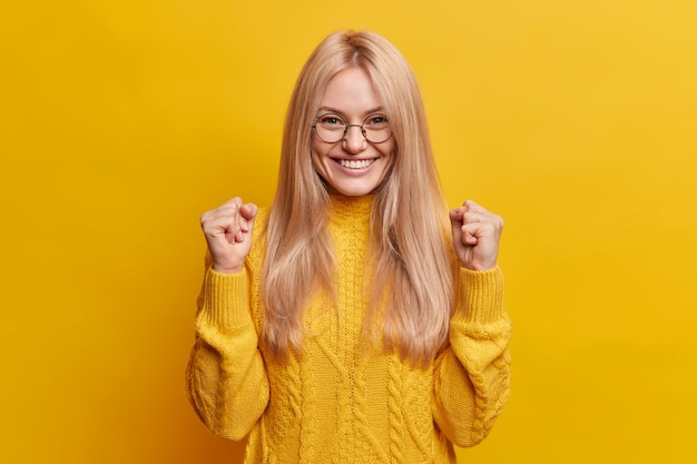 Feliz y alegre mujer rubia aprieta los puños y celebra el buen resultado sonríe ampliamente estando en alto espíritu hace gesto de triunfo usa anteojos y suéter de invierno