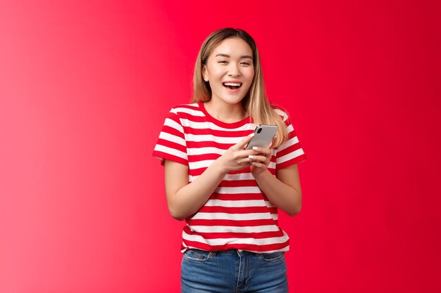 Feliz alegre niña asiática rubia riendo con teléfono inteligente dispositivo de retención de niña sonriendo ampliamente mirada vino vino ...