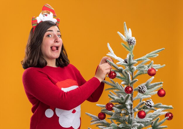 Feliz y alegre niña en suéter de Navidad con diadema divertida de pie junto a un árbol de Navidad colgando bolas de Navidad sobre la pared naranja