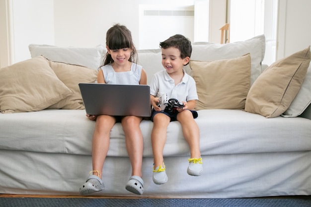 Feliz alegre niño y niña sentados en el sofá en casa, usando la computadora portátil, viendo videos, películas de dibujos animados o películas divertidas.