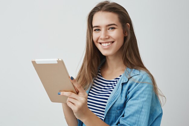 Feliz amigable hermosa asistente femenina sonriendo ampliamente sosteniendo tableta digital, posando alegremente, satisfecho de lo fácil que es dibujar usando un gadget