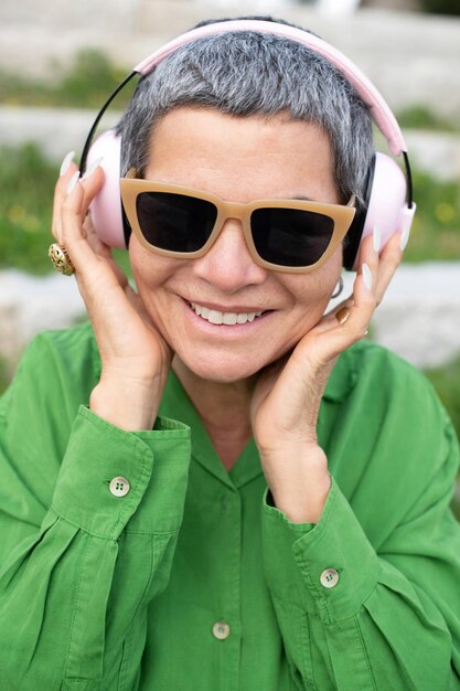Feliz anciana escuchando música en el parque. Modelo femenino con cabello gris corto en ropa brillante y grandes auriculares. Ocio, actividad, concepto de tecnología.