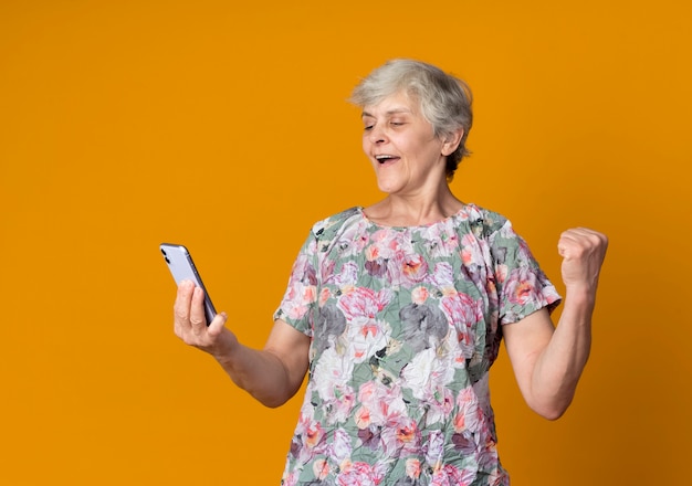 Foto gratuita feliz anciana levanta el puño y mira el teléfono aislado en la pared naranja