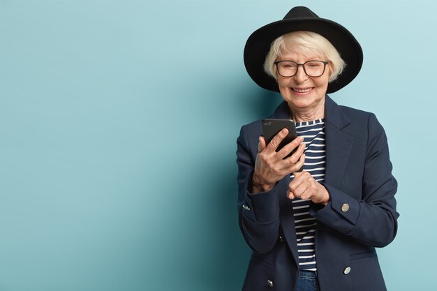 Feliz anciana recibe buenas noticias en el teléfono inteligente, escribe comentarios, usa un sombrero negro de moda