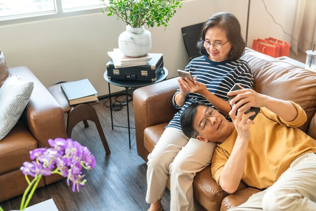 Feliz anciano amante asiático pareja sosteniendo teléfono inteligente mirando la pantalla del teléfono celular riendo casual relajante sentarse en el sofá juntos sonriendo ancianos abuelos maduros familia abrazando el estilo de vida