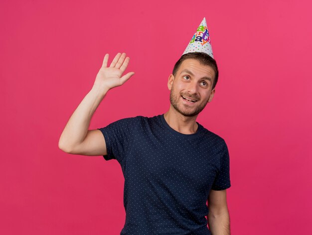 Foto gratuita feliz apuesto hombre caucásico con gorro de cumpleaños se encuentra con la mano levantada aislada sobre fondo rosa con espacio de copia