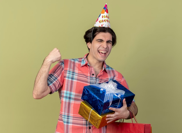 Feliz apuesto hombre caucásico con gorro de cumpleaños mantiene el puño sostiene cajas de regalo y bolsa de papel