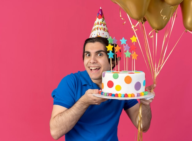 Foto gratuita feliz apuesto hombre caucásico con gorro de cumpleaños sostiene globos de helio y pastel de cumpleaños