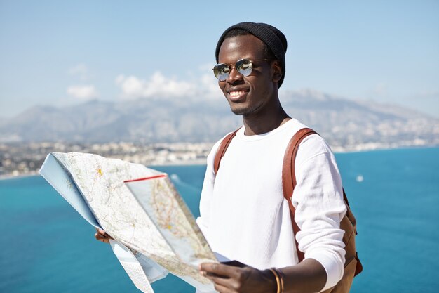 Feliz apuesto joven viajero de piel oscura de pie en la cima de la montaña con un mapa de papel sobre el vasto océano y la ciudad turística, con una mirada alegre mientras viaja por el mundo en compañía de amigos