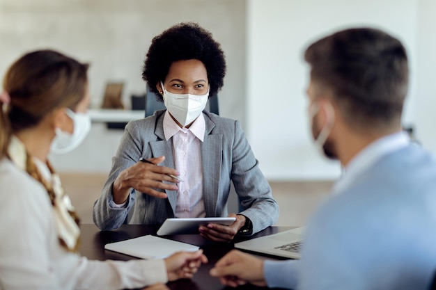 Feliz asesor financiero afroamericano consultando a sus clientes mientras usa mascarilla protectora en una reunión