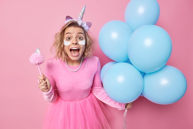 Feliz asombrada joven europea exclama en voz alta aplica parches de belleza debajo de los ojos usa vestido sostiene un montón de globos azules inflados aislados sobre fondo rosa Concepto de ocasión festiva