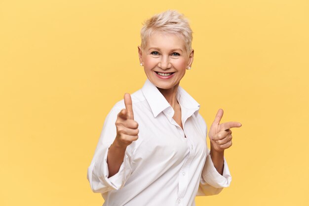 Feliz atractiva mujer de cincuenta años con elegante camisa blanca apuntando con el dedo índice y sonriendo, eligiéndote para bailar con ella, mirando con amplia sonrisa radiante. Lenguaje corporal