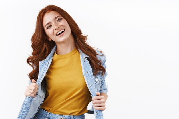 Feliz atractiva mujer pelirroja con estilo en chaqueta de mezclilla, mostrando la camiseta amarilla, inclinar la cabeza y sonriendo satisfecho, sintiéndose despreocupado y alegre, pared blanca