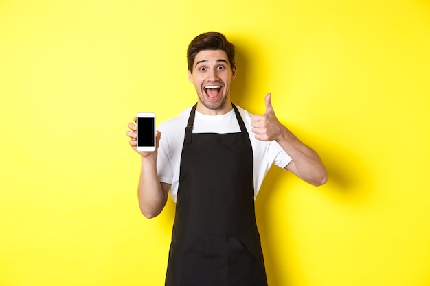 Feliz barista en delantal negro mostrando la pantalla del teléfono inteligente, hacer pulgar hacia arriba, recomendando la aplicación de café, de pie sobre fondo amarillo.
