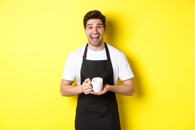 Feliz barista en delantal negro sosteniendo la taza de café, riendo y de pie sobre fondo amarillo.