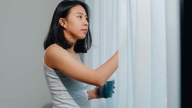 Feliz bella mujer asiática sonriendo y bebiendo una taza de café o té cerca de la ventana en el dormitorio. La muchacha latina joven abre las cortinas y se relaja en mañana. Señora de estilo de vida en casa.