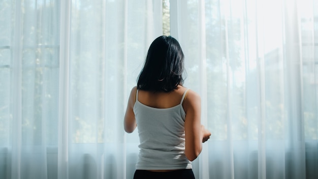 Feliz bella mujer asiática sonriendo y bebiendo una taza de café o té cerca de la ventana en el dormitorio. La muchacha latina joven abre las cortinas y se relaja en mañana. Señora de estilo de vida en casa.