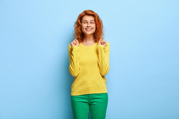 Feliz bonita mujer jengibre aprieta los puños, se siente muy bien después de ganar la competencia, vestida de manera informal